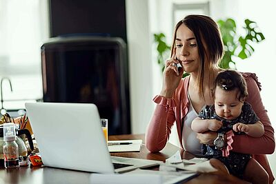 Frau telefoniert mit Baby auf dem Arm vor Laptop