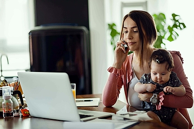 Frau telefoniert mit Baby auf dem Arm vor Laptop